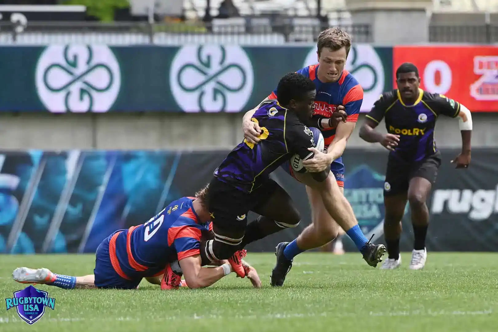 Two rugby players in blue and red try to tackle a player in a black and purple uniform during a match on a grassy field.