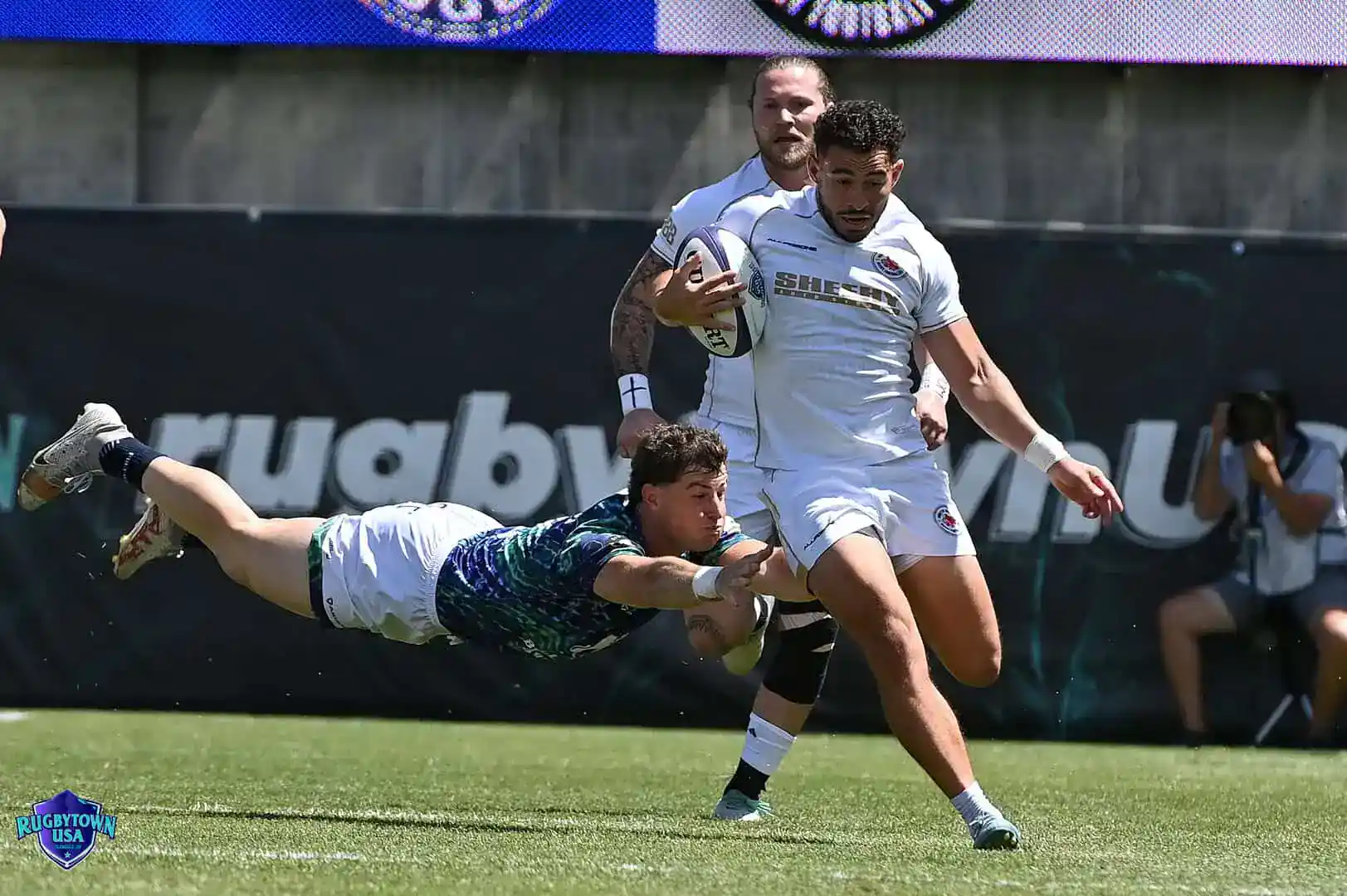 A rugby player in white runs with the ball while a player in blue dives to tackle him; another player follows behind on a grassy field.