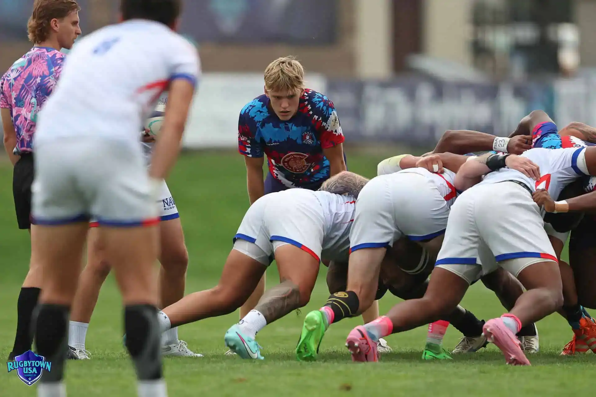 Rugby players engage in a scrum on a grassy field, with one player in a blue jersey observing closely as others in white uniforms push against opponents.