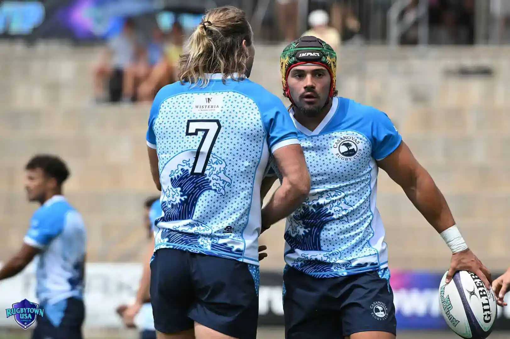 Two rugby players in blue and white jerseys, one holding a ball, appear to be communicating during a match on an outdoor field.