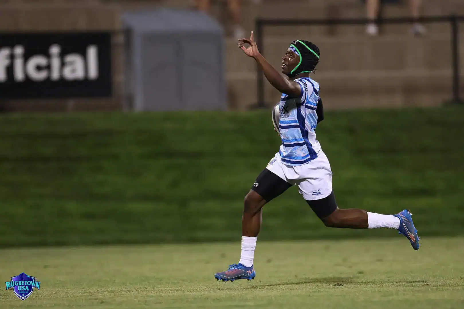 A rugby player in a blue and white jersey runs on the field with one arm raised, celebrating during a nighttime match.